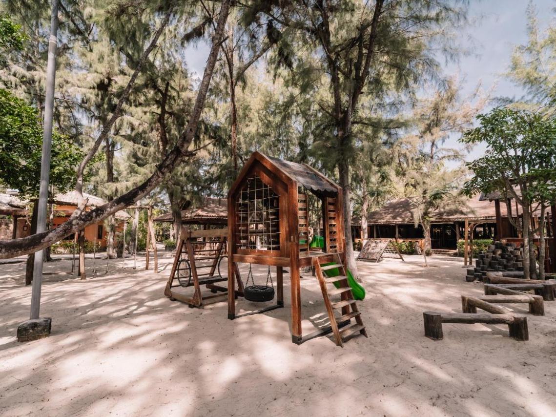 Sand playground under casuarina trees with a wooden climbing house, ladder and slide
