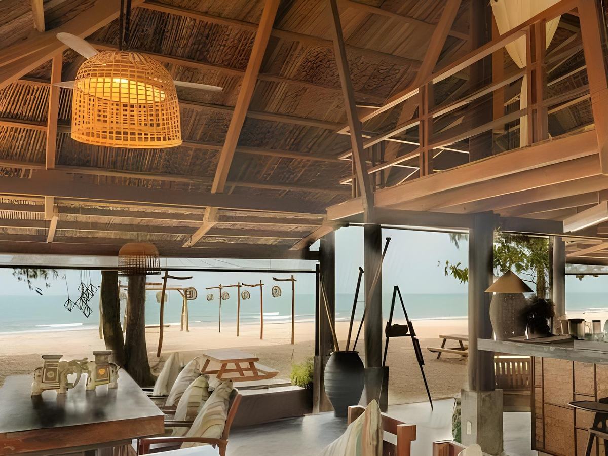 Open-sided beach bar under a thatched and timber roof with tables, cushions and the sand and sea beyond