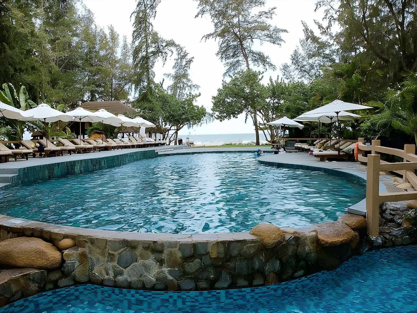 Curving pool with a stone edge and a small wooden bridge, white umbrellas and loungers, casuarina trees and the sea on the horizon