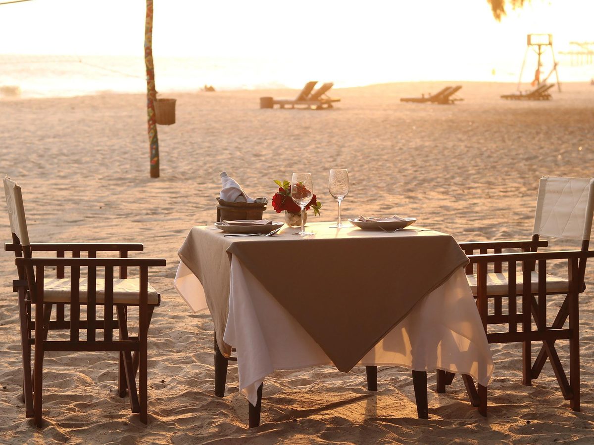 Table for two on the sand at sunset with a white cloth and two wooden folding chairs, loungers and the beach behind