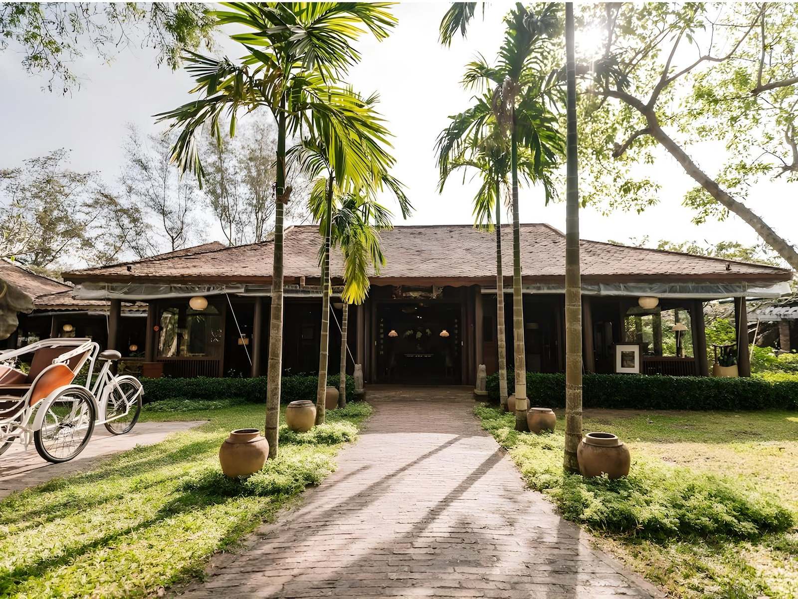 Paved path between areca palms leading to an open timber pavilion with a tiled roof, bicycles parked on the left and clay jars on the lawn