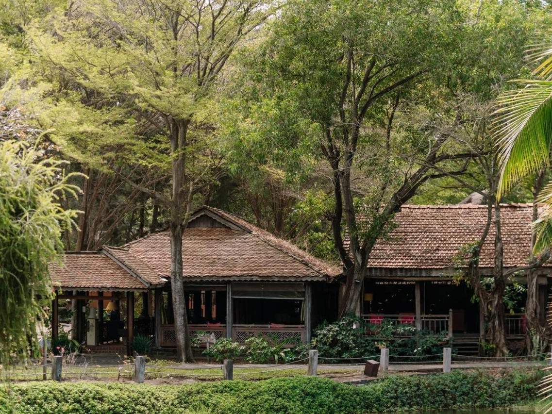 Low timber house with a clay-tile roof under tall trees, a clipped hedge and lawn in front