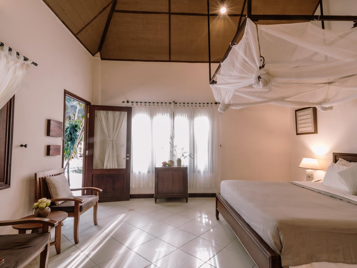 Bungalow bedroom with a four-poster bed and white net, wooden armchairs, tiled floor and a high thatched ceiling