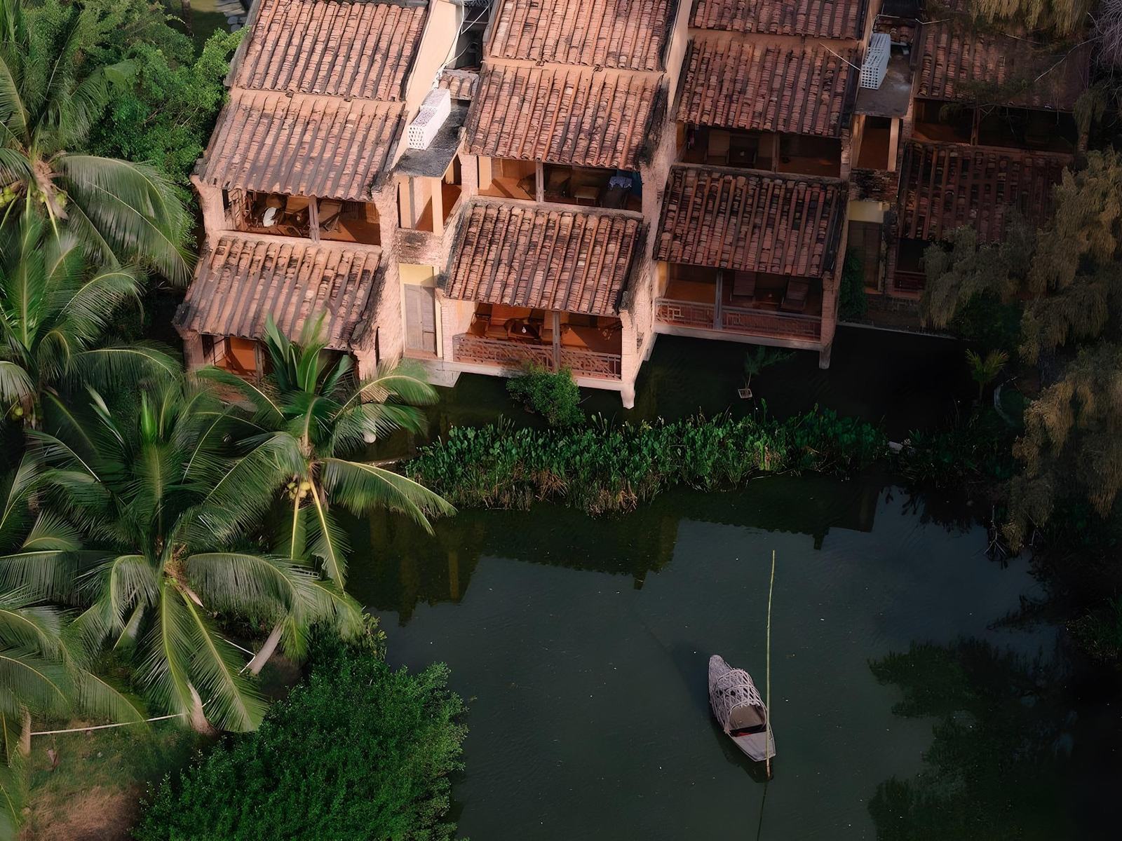 Aerial view of two-storey red-brick houses with clay-tile roofs beside a dark lagoon, coconut palms in front and a small wooden boat on the water