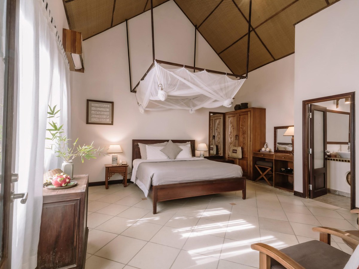 Bright room under a high thatched ceiling, double bed with a white mosquito net, tiled floor and carved wooden cabinet