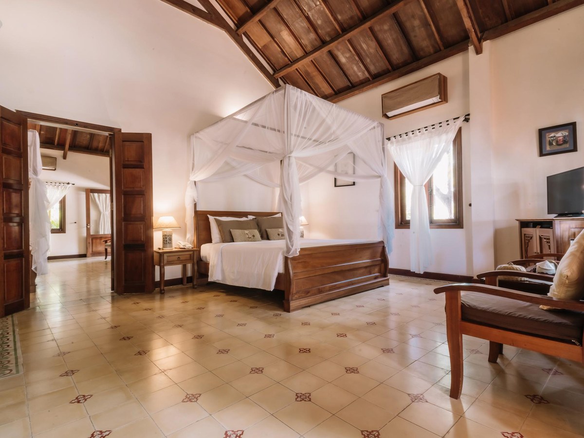 Spacious bedroom with a four-poster bed, cream tiled floor, wooden sofa and doors open to a bright terrace