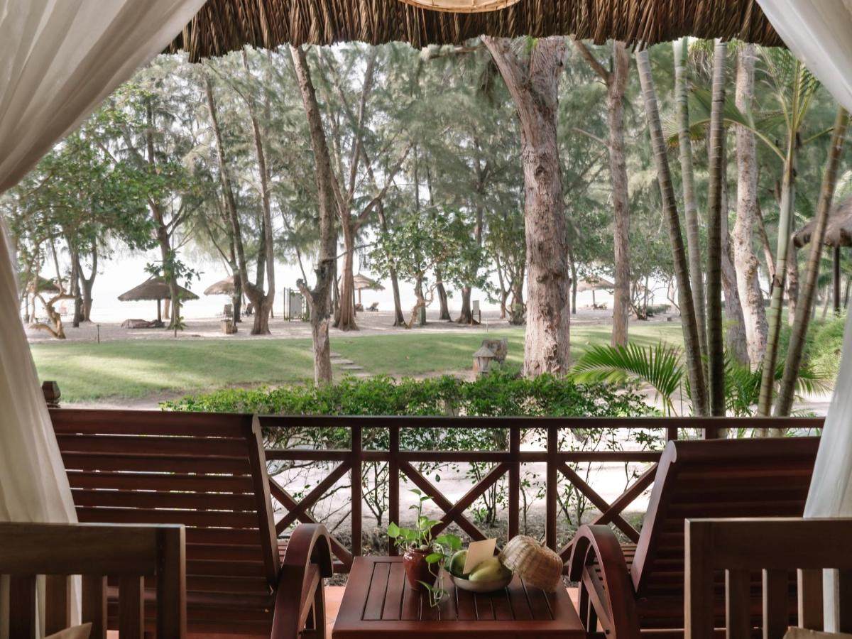Wooden veranda with two armchairs, white curtains pulled back, casuarina trees and lawn leading to the beach