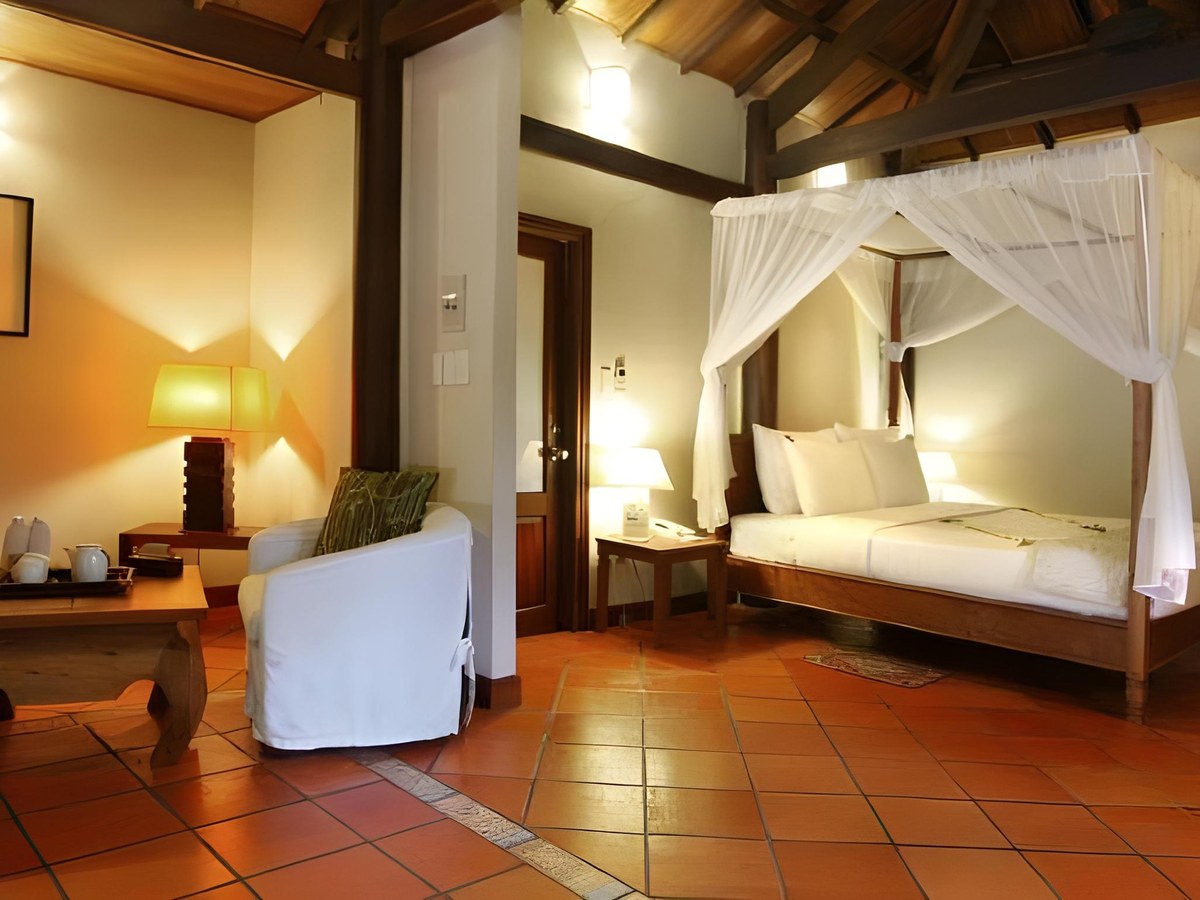 Bedroom with terracotta floor tiles, white four-poster bed, armchair and warm lamps under a sloping wooden ceiling