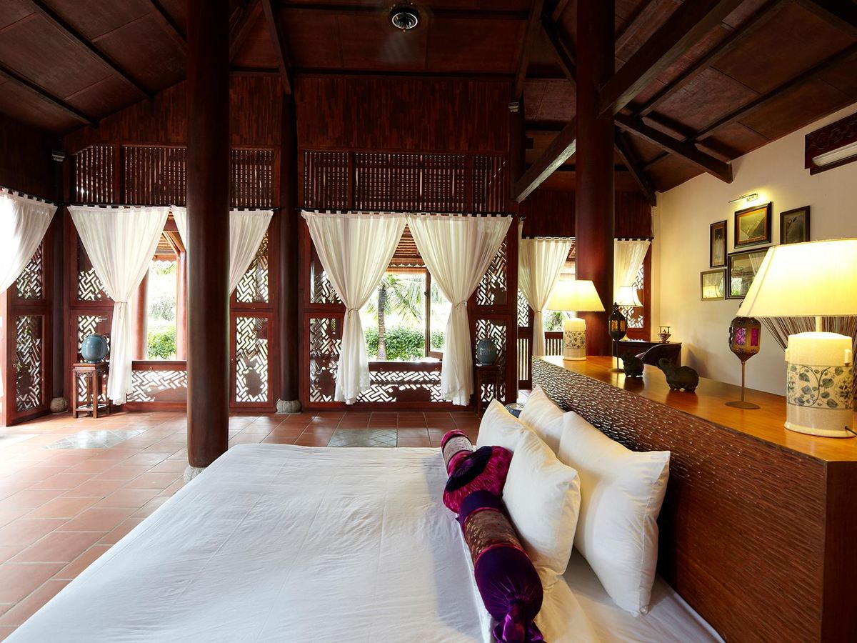 Bedroom with dark wooden pillars, carved lattice windows, a white bed with purple cushions and a table lamp