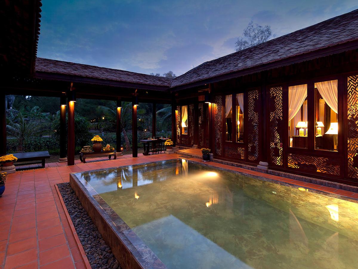 Courtyard pool at dusk enclosed by dark wooden pavilions with lit windows and a tiled terrace