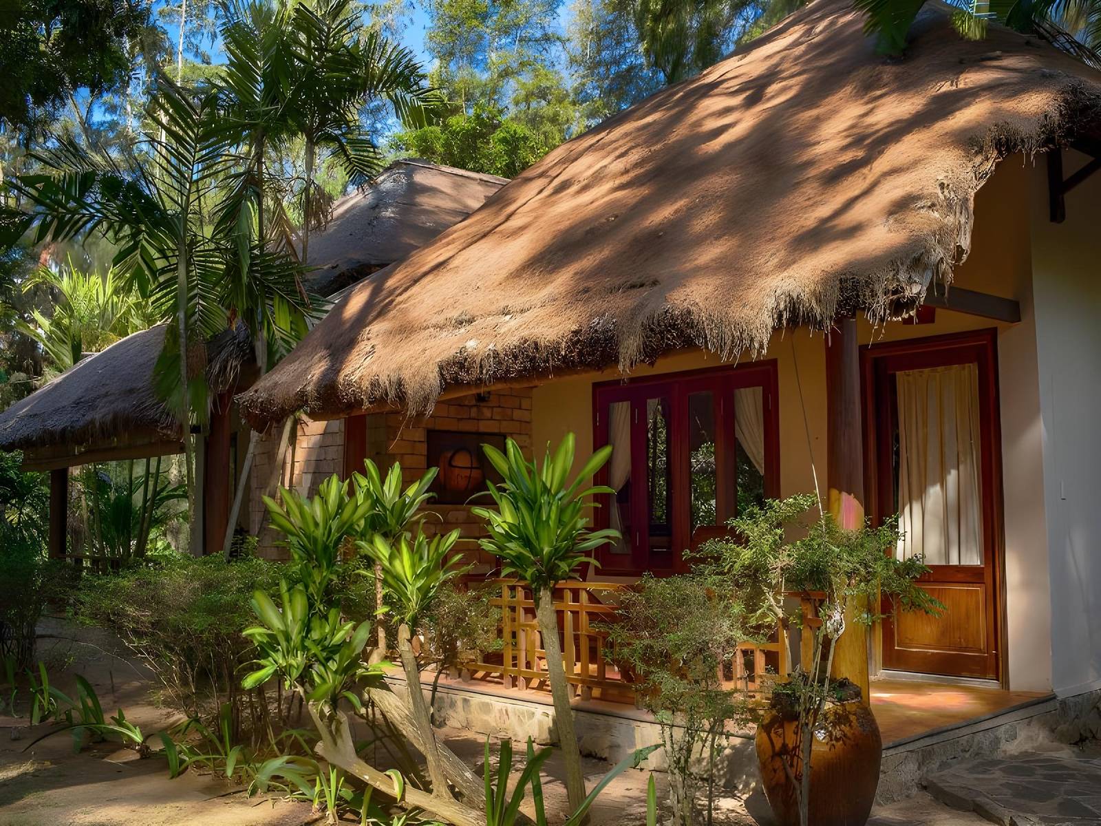 Thatched bungalow with ochre walls and red-brown wooden doors behind palms and tropical shrubs in bright sun