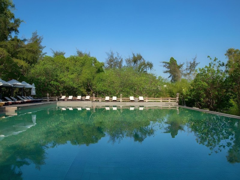 Long straight pool reflecting tall casuarina trees and a blue sky, white umbrellas along the far edge