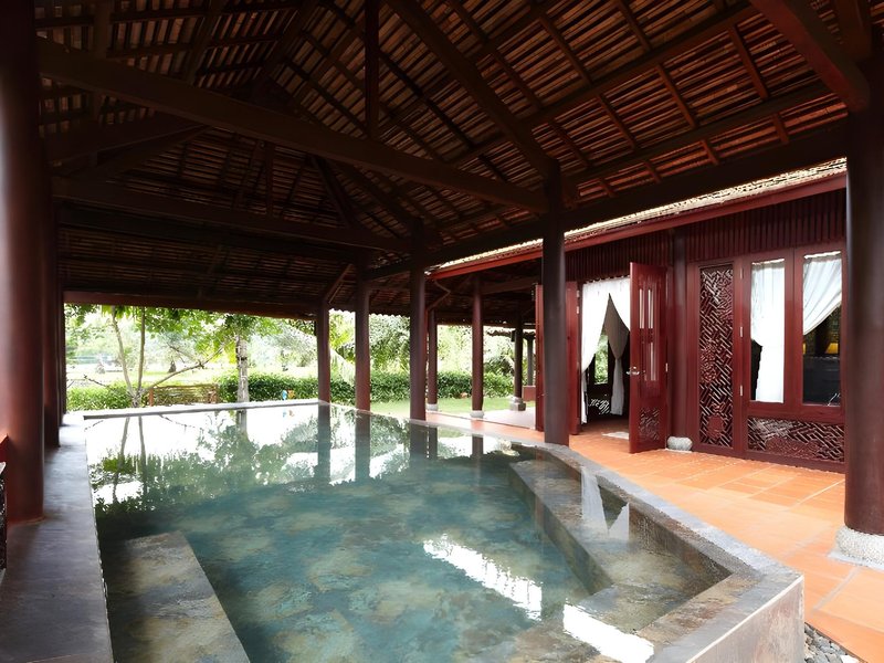Private pool under a deep wooden veranda with dark red carved pillars and lattice doors, terracotta floor tiles